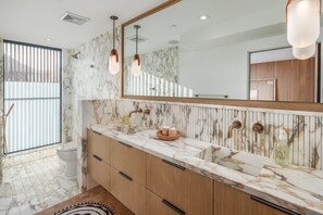 Une salle de bain avec un lavabo en marbre, des meubles en bois, un grand miroir et une fenêtre avec des stores.