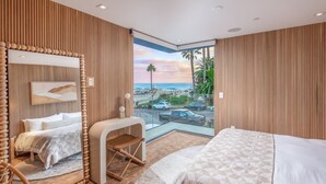 Habitación de hotel moderna con una cama grande, un área de descanso, una mesita y un ventanal con vistas a la playa y palmeras.