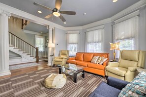 A spacious living room with a modern orange sofa, a glass coffee table, and a beige armchair. A ceiling fan and a staircase are visible in the background. - Elegant Raleigh Home w/ Porch, Walk Downtown! (Raleigh)