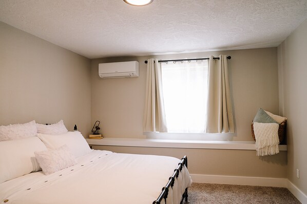 A bedroom with a bed, a window with curtains, a bedside table with a lamp, and a wall-mounted air conditioner.