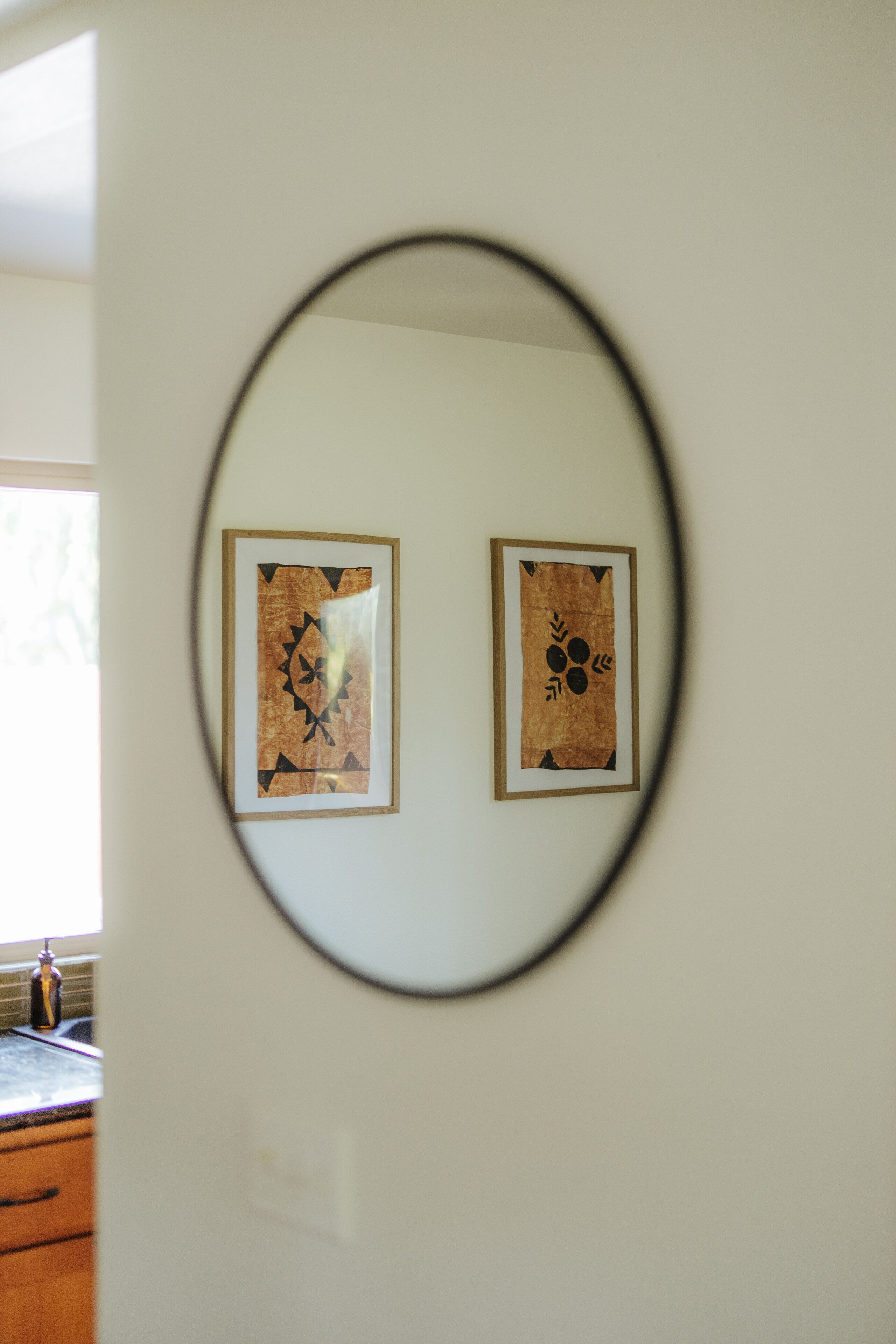 Two framed artworks on a wall, a kitchen area with wooden cabinets and a window, and a round mirror reflecting part of the room.