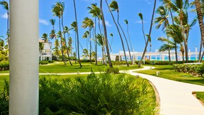 A well-maintained garden with palm trees and a pathway leading to a building with a pool. - Playa Turquesa I-202 Premier Beachfront Ocean View 85 mbps wifi (Punta Cana)