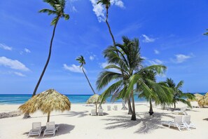Ein Strand mit weißen Liegestühlen, Palmen und Reetdachschirmen.