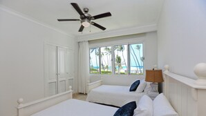 A hotel room with two beds, a ceiling fan, a window with a view of palm trees and the ocean, and a lamp on the nightstand. - Playa Turquesa I-202 Premier Beachfront Ocean View 85 mbps wifi (Punta Cana)