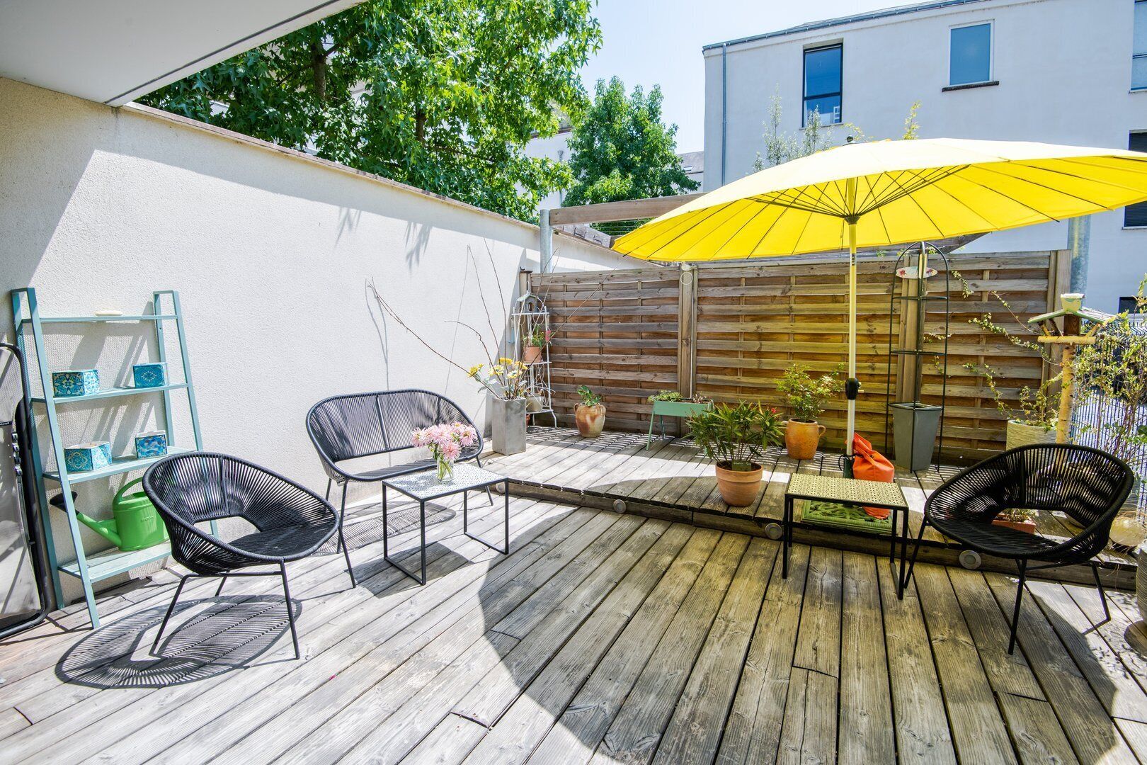 Una terraza de madera con mobiliario exterior, una sombrilla amarilla y plantas en macetas.