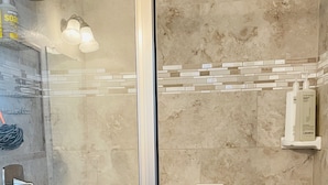 Area doccia con porta in vetro, soffione a mano, dispenser di sapone e un piccolo ripiano con articoli da toilette.