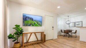 Uma sala de estar moderna com piso de madeira, uma televisão de tela plana grande fixada na parede, uma área de jantar com mesa e cadeiras, e um vaso com planta.