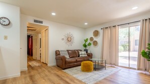 Uma sala de estar moderna com um sofá de couro marrom, uma mesinha redonda pequena e um banquinho amarelo. Há uma janela grande com cortinas, um relógio de parede e arte decorativa na parede.