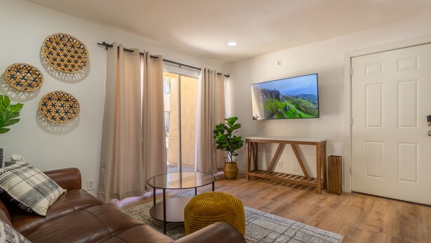Uma sala de estar com sofá de couro marrom, mesa de centro de vidro, um banquinho amarelo, uma TV sobre um móvel de madeira e uma porta de vidro deslizante que dá para uma varanda.