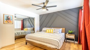 Quarto com cama grande, ventilador de teto, mesa de cabeceira com planta e espelho na parede.