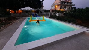 Une piscine avec un toboggan et des jeux gonflables, entourée d’une zone pavée et bordée de bâtiments en arrière-plan.