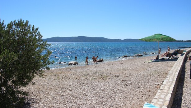 A pebble beach with a clear blue sea, a green hill in the background, and a few people enjoying the water. - NEW Holiday Apartment,A4+2, Family house - OPG Rudan, garden pool, terase, wi-fi (Radonić)