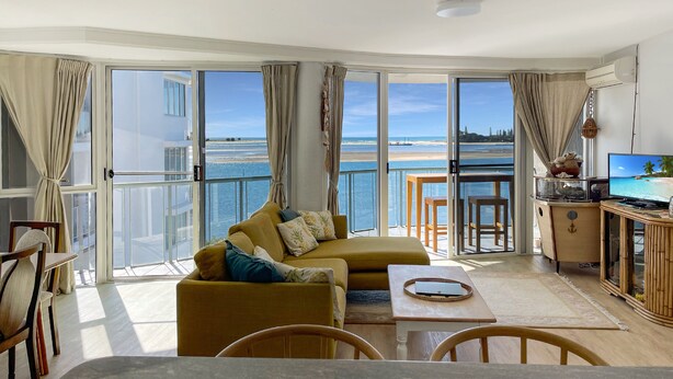 Una sala de estar moderna con sofá, mesa de comedor y televisor. Amplias ventanas ofrecen vista al mar y un balcón.
