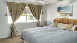 Un dormitorio con una cama grande, cortinas color beige, un cabecero de madera y un cuadro con motivos marinos en la pared.