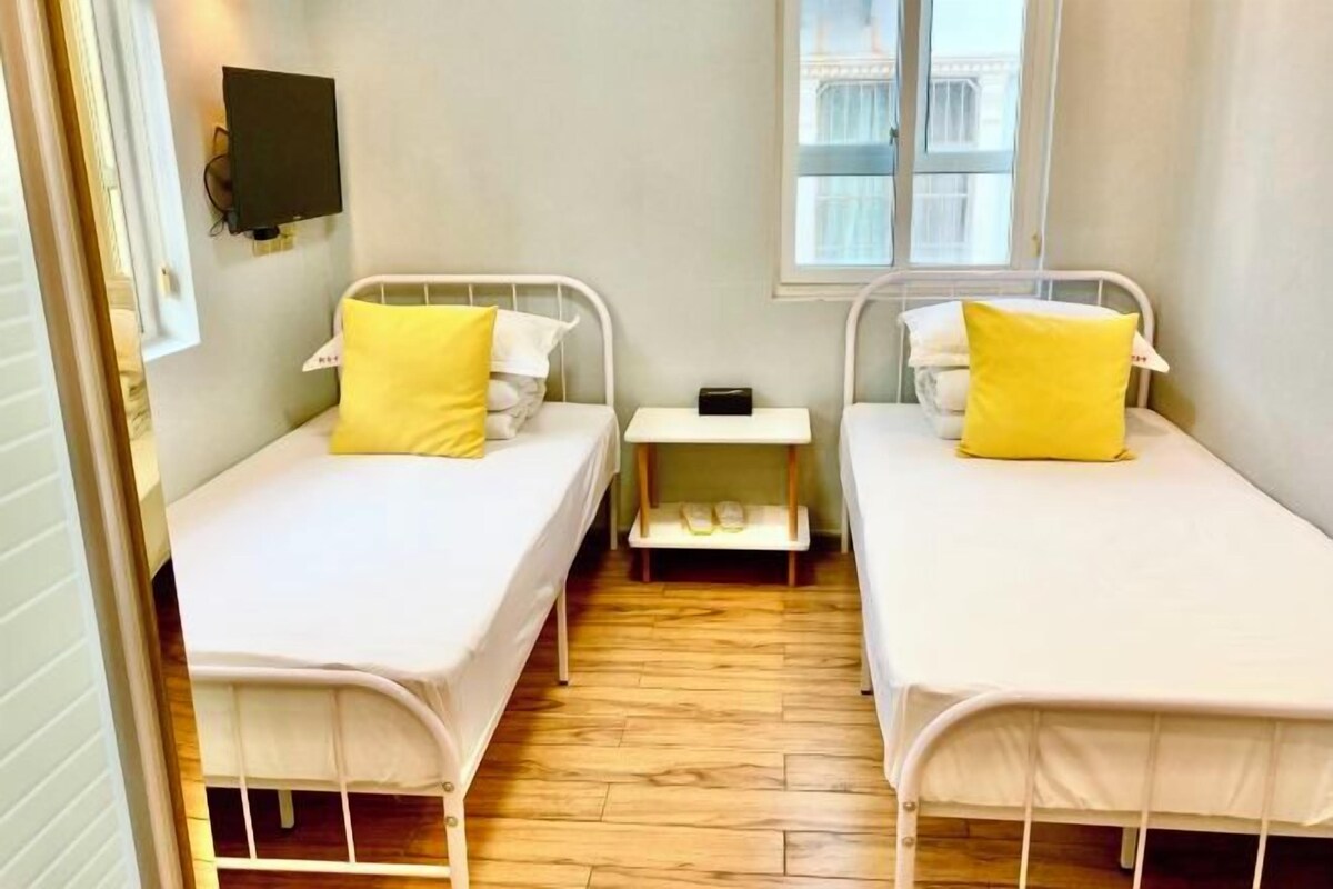 A small, clean room with two single beds, each with a white mattress and a yellow pillow. There is a small bedside table with a lamp and a power outlet. A window provides natural light, and a flat-screen TV is mounted on the wall.