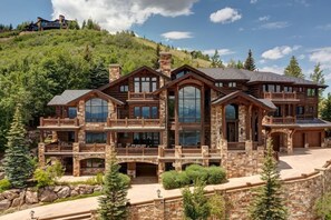 A large, multi-story log cabin with multiple balconies and large windows, surrounded by a stone retaining wall and lush greenery. - Abode at Chateau Jordan, ski-in/ski-out, hot tub, gated, Deer Valley (Heber City)