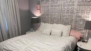 Een net opgemaakt bed met een witte deken en kussens, een nachtkastje met een lamp en een decoratief hoofdeinde met gedetailleerde patronen.
