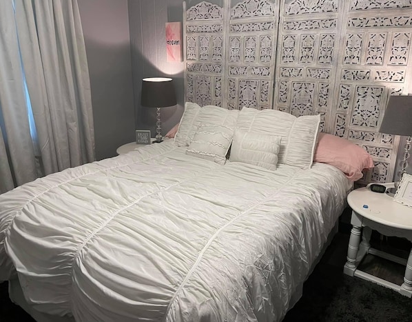 Una cama bien hecha con edredón blanco y almohadas, una mesita de noche con lámpara, y un cabecero decorativo con diseños intrincados.