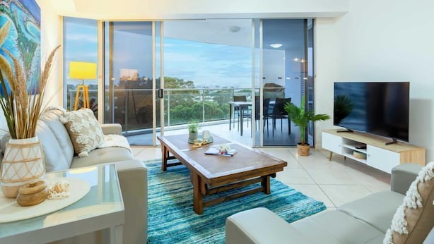 A modern living room with a large TV, a sofa, a coffee table, and a balcony with a view.