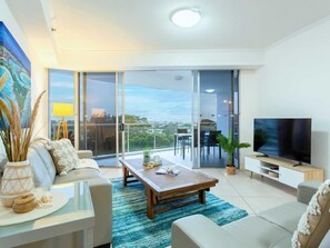 A modern living room with a large TV, a sofa, a coffee table, and a balcony with a view.