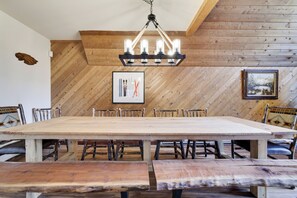 Une grande table à manger en bois, avec des bancs, entourée de chaises, dans une pièce aux murs en bois et agrémentée d’un luminaire suspendu.