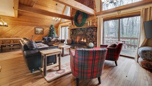 A cozy cabin interior with a stone fireplace, wooden beams, and a large window overlooking a deck. - Mountain chalet with 6 BR, hot tub, game room and fire pit. (Lake Harmony)