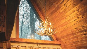 A cozy cabin interior with a wooden ceiling, a plaid sofa, a fireplace, and a large window overlooking a forest. - Mountain chalet with 6 BR, hot tub, game room and fire pit. (Lake Harmony)