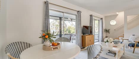 Una sala de estar moderna con una mesa redonda blanca, sillas estampadas, un televisor y vistas amplias por grandes ventanales.