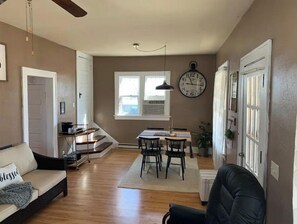 A living room with a sofa, a dining table with chairs, a clock on the wall, and a staircase leading to another room. - Entire house for fishing, hunting, or passing by! (Fairfax)