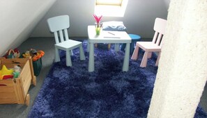 Una habitación con alfombra azul, dos sillas blancas, una mesa pequeña con un florero rosa y una ventana.