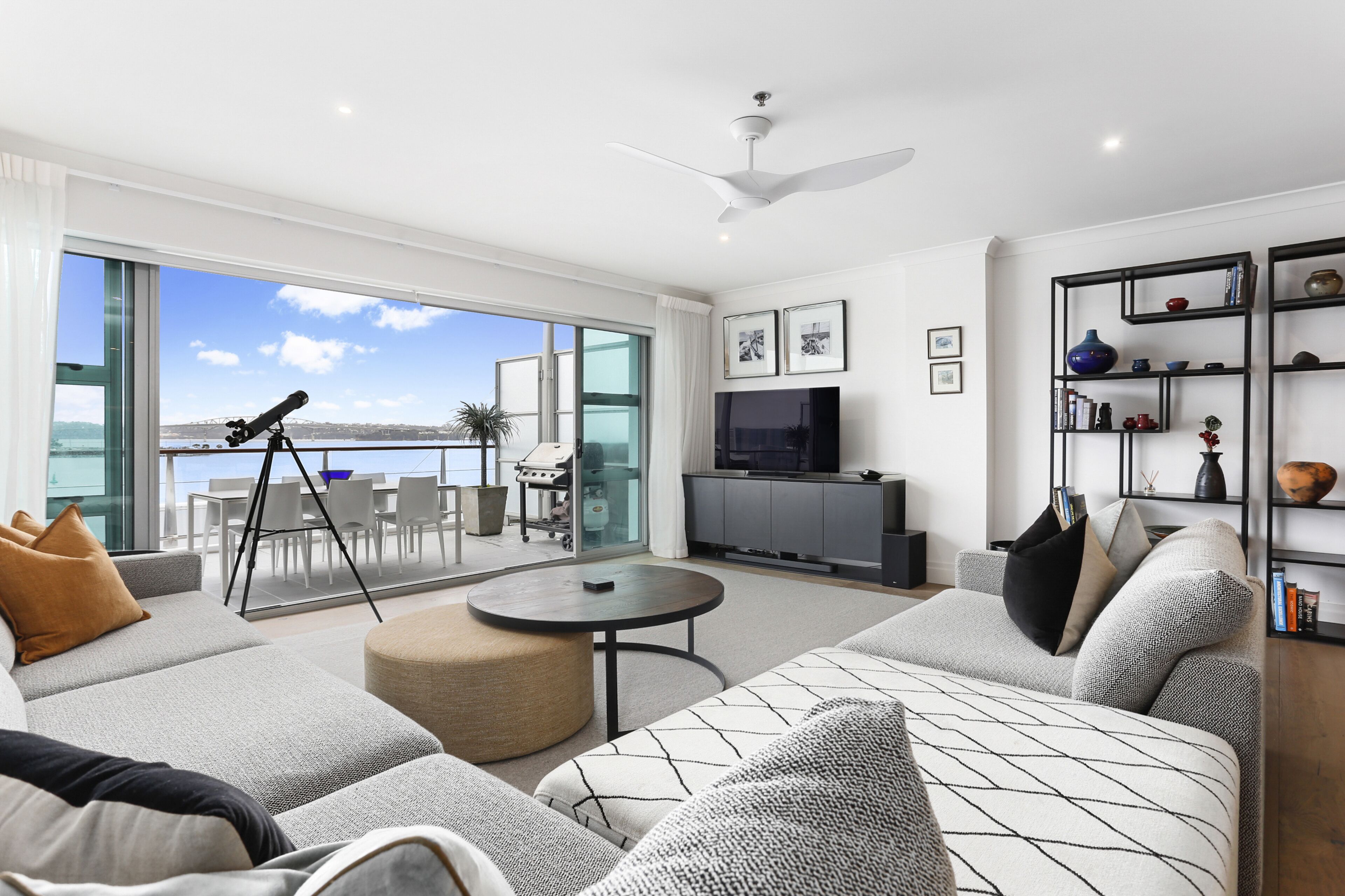 A modern living room with a large sofa, a round coffee table, and a telescope on a balcony overlooking a body of water.