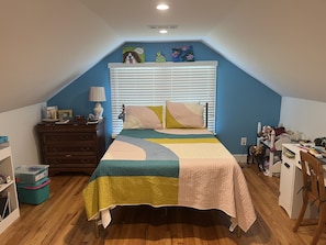 A bedroom with a bed, a dresser, a desk, and a window with blinds.