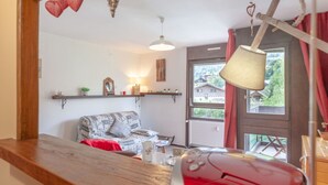 Un salon chaleureux avec un canapé, une table en bois et un fauteuil rouge. Une fenêtre offre une vue sur les maisons et les arbres.