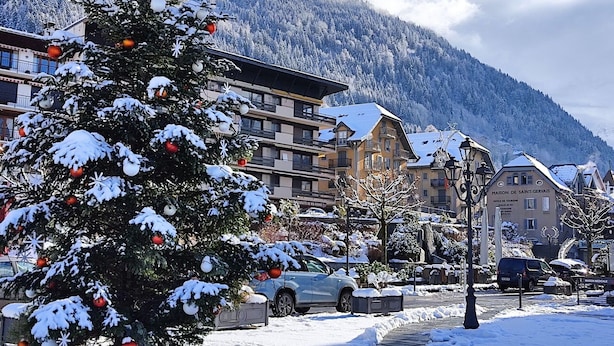 Un sapin de Noël recouvert de neige, installé sur une place de ville, avec des bâtiments et des montagnes en arrière-plan.
