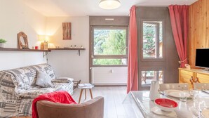 Un salon chaleureux avec un canapé, une table à manger dressée pour deux personnes et une grande fenêtre donnant sur un paysage verdoyant.