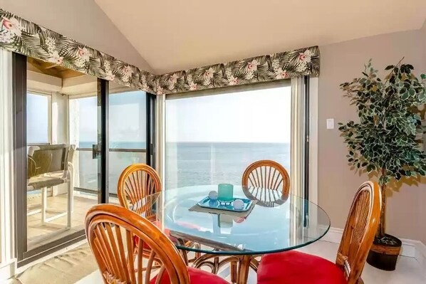Una mesa de vidrio con cubierta de cristal y cuatro sillas de madera, una mesa de vidrio con una vela azul y una bandeja, y vistas al mar a través de amplias ventanas.