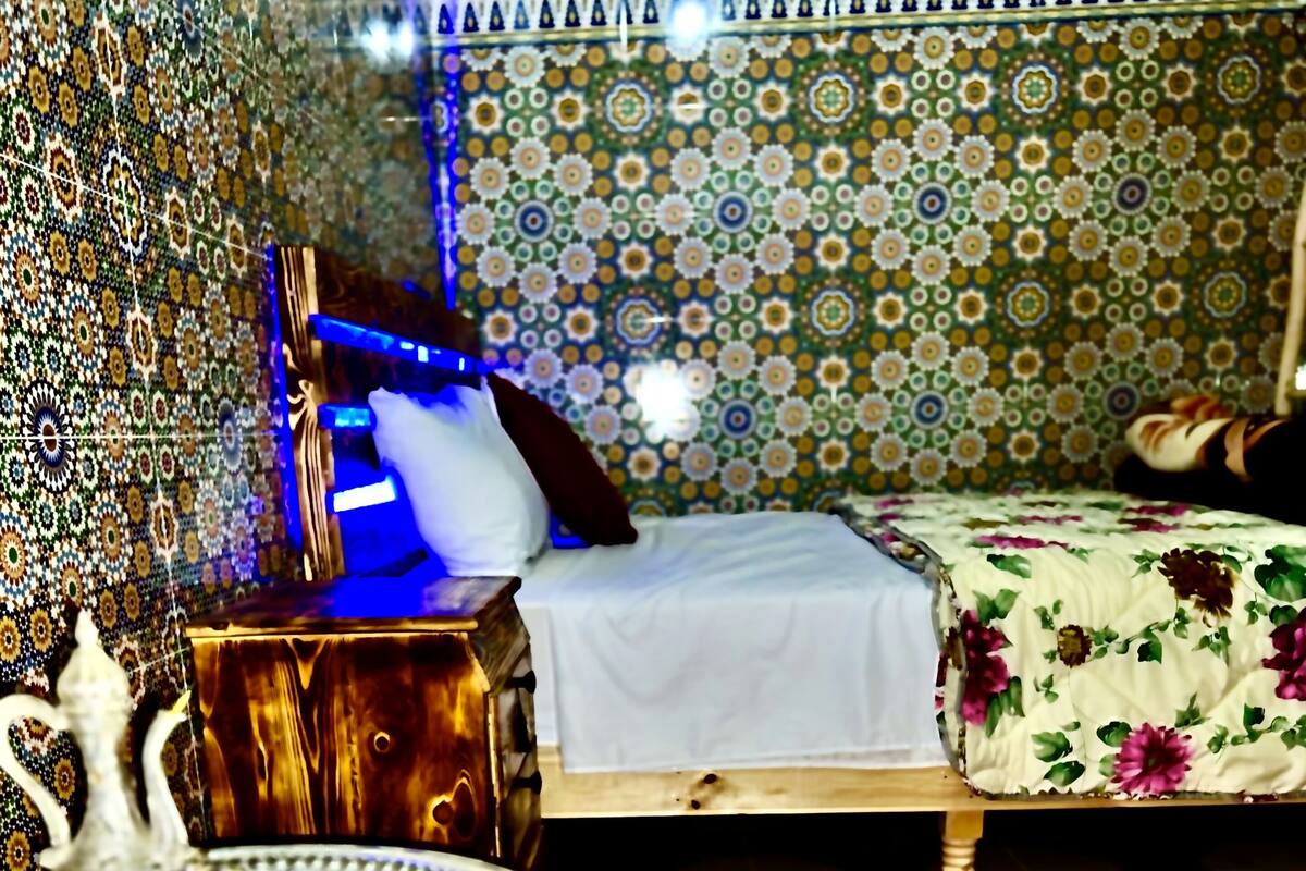 Una habitación con pared estampada, cama con colcha floral, una mesita con adorno y un taburete tejido y colorido.
