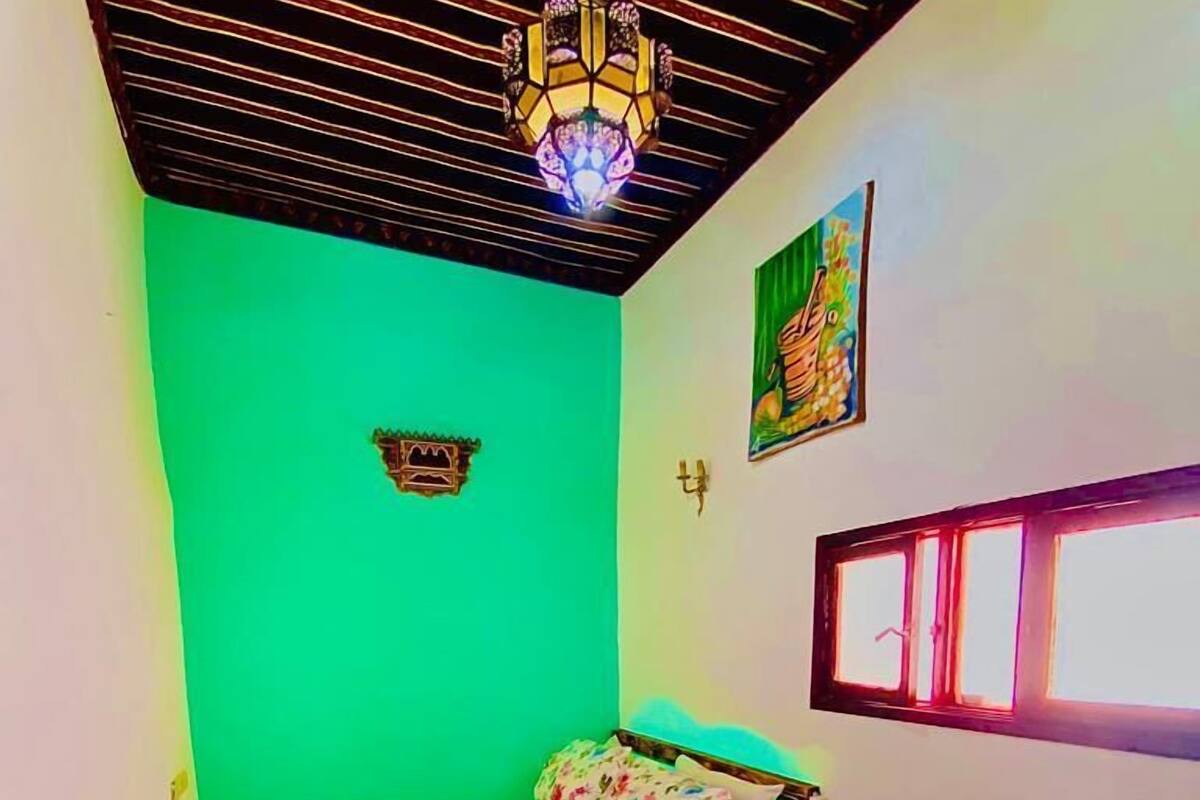 Una habitación con pared verde, cama con ropa de cama floral, cabecero de madera, mesita de noche pequeña, lámpara, un cuadro, ventana y una silla colgante.