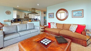 Ein Wohnzimmer mit grauem Sofa, einem braunen Lederhocker und einem braunen Sofa mit roten Kissen. An der Wand hängt ein runder Spiegel und zwei gerahmte Bilder.
