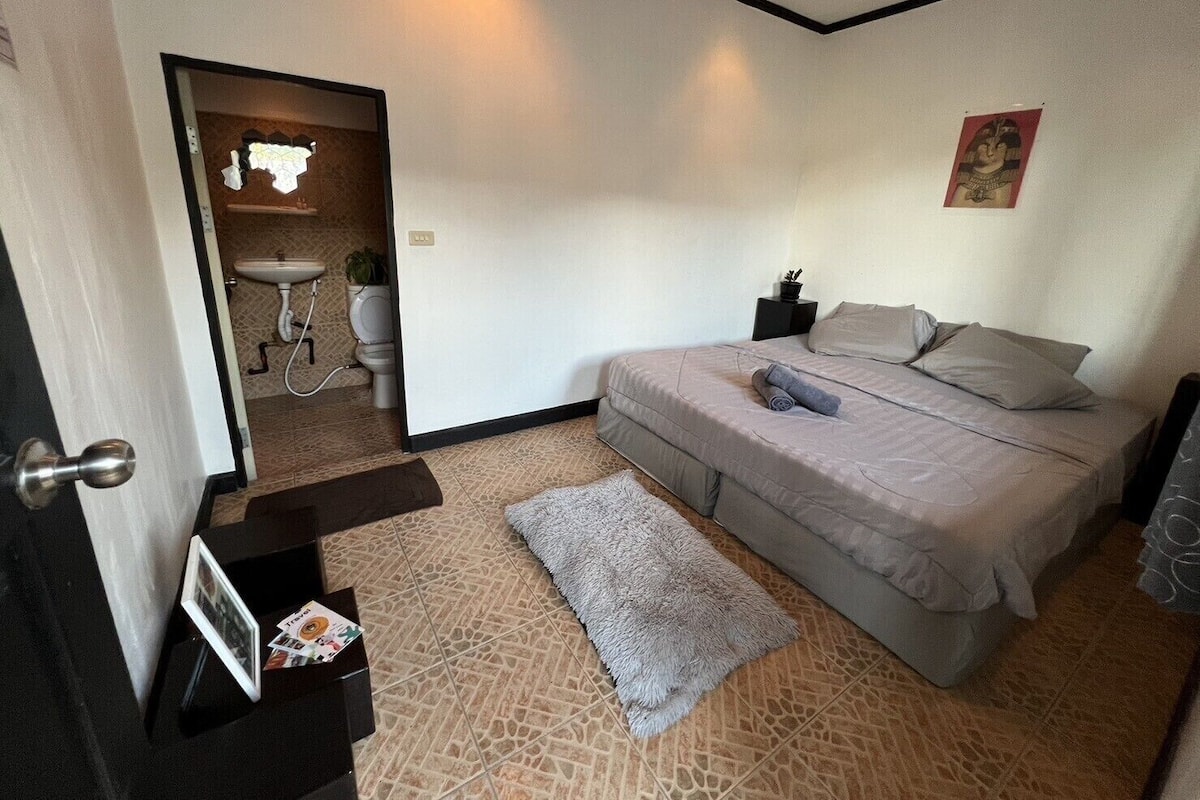 Un dormitorio con cama, mesita de noche, estante en la pared, una puerta y un baño visible a través de una puerta abierta.