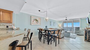 A spacious living area with a dining table, chairs, and a sofa. There is a kitchen area with wooden cabinets and a countertop. - ⛱️SummerChase 904⛱️ indoor pool, hot tub, kiddie pool, gym, and beach towels (Orange Beach)