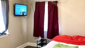 A hotel room with a bed, a TV mounted on the wall, a small table with a bottle, and a window with curtains. - The Ugly House (Houston)