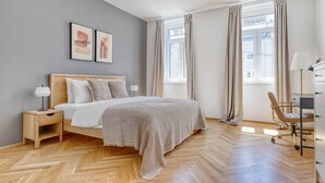 Et moderne soveværelse med gulv i træ i fiskebensmønster, en seng med gråt og hvidt stribet dyne, et natbord med lampe, et skrivebord med stol og to indrammede kunstværker på væggen.