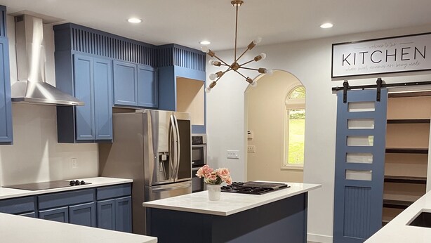 A modern kitchen with blue cabinetry, a central island, stainless steel appliances, and a 'KITCHEN' sign.