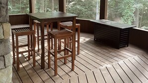 A wooden deck with a table and chairs, large windows, and a stone wall.