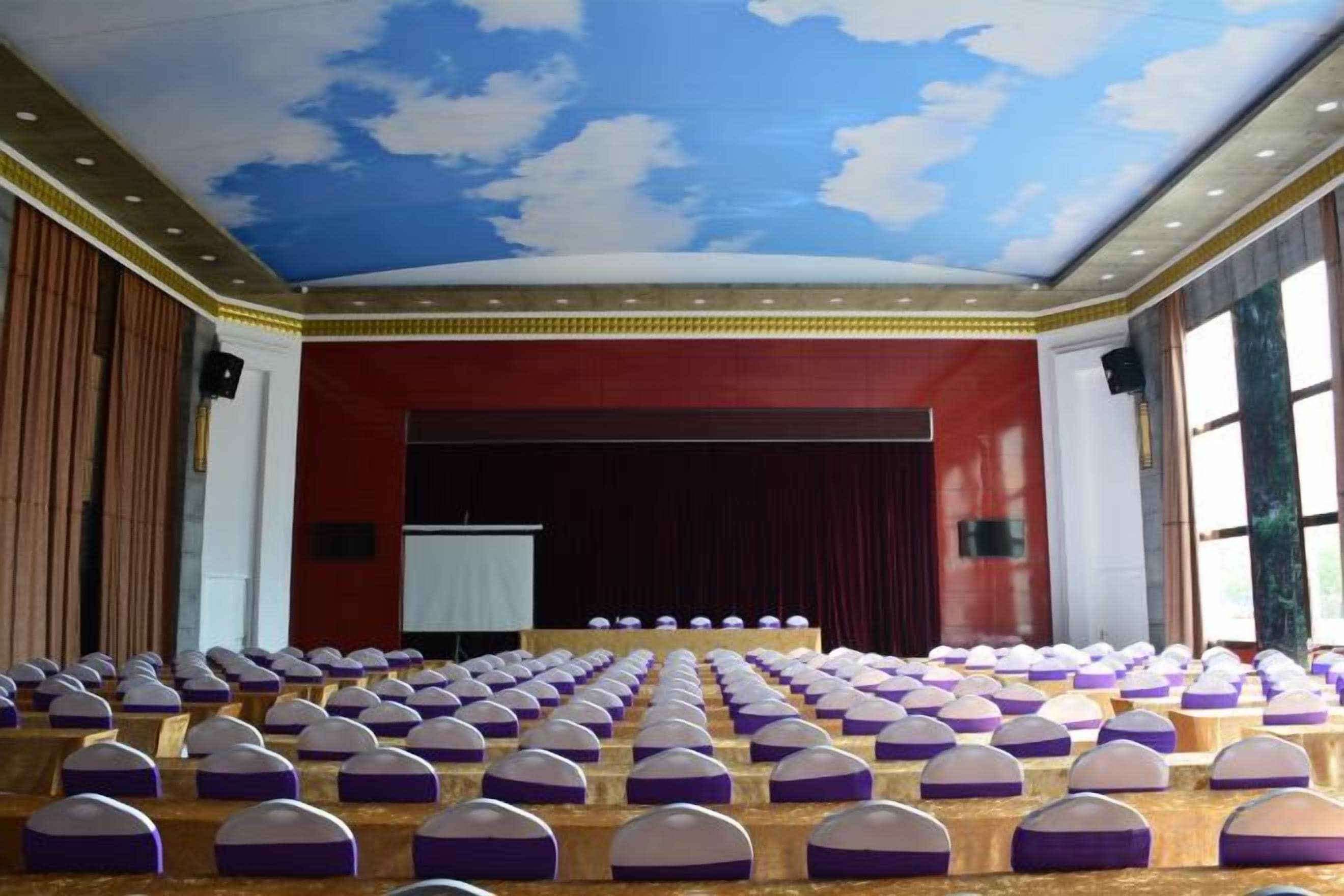 A large hall with rows of seats and a stage, a red wall, and a blue ceiling with cloud patterns.