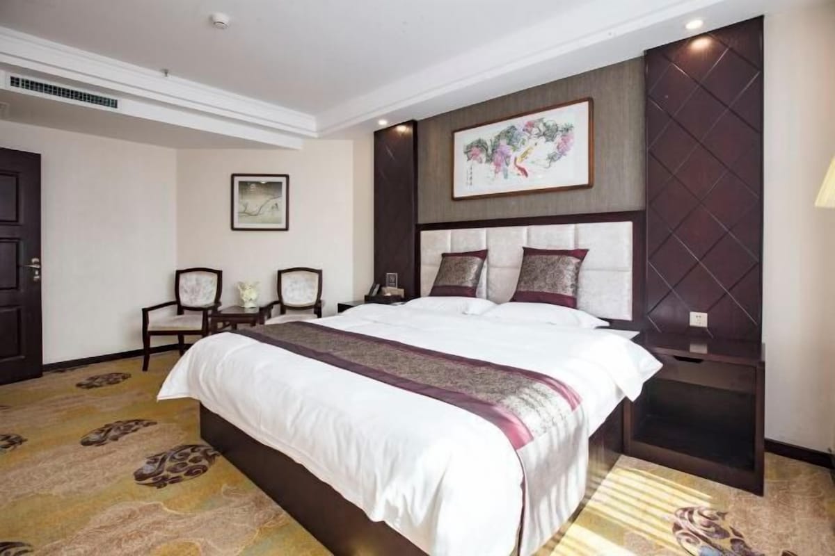 A hotel room with a large bed, a headboard with a decorative panel, a framed artwork above the bed, and a carpet with a pattern.