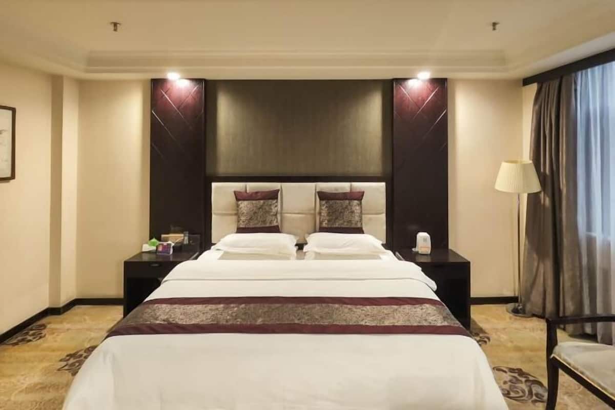 A hotel room with a large bed, two bedside tables, a nightstand, a lamp, and a window with curtains.