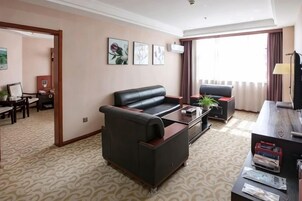 Habitación de hotel con sofá, mesa de centro, televisor y escritorio. Se aprecia una ventana con cortinas, una puerta que da a otra habitación y una alfombra estampada.