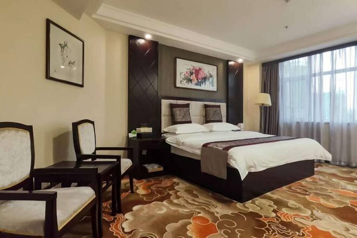 A hotel room with a bed, a desk, two chairs, a nightstand, a lamp, and a window with curtains.